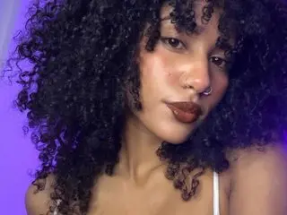 CinthiaWalker poses with her curly dark hair framing her face, showing her bare shoulders and neck against a purple background.