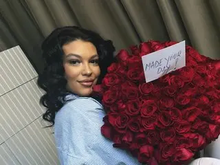 ClaraaPasssion poses with long dark hair, wearing a light blue button-up shirt while leaning against a wall next to a large bouquet of red roses.