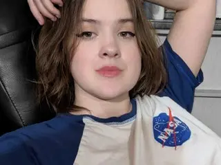 ClickGirl has shoulder-length brown hair and wears a white and blue t-shirt while sitting back in a black chair.