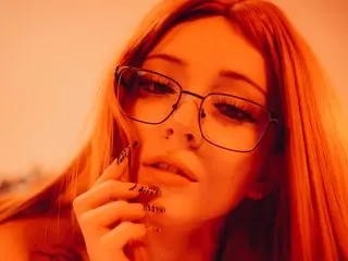 Cloefive poses with her hand near her face, looking at the camera with long hair and glasses against a warm, orange-toned background.
