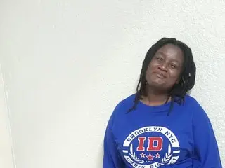 CocoBerryZA wearing a blue Brooklyn NYC sweatshirt, looking at the camera with her hair down against a plain white wall.