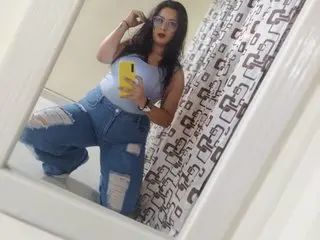 ColsexyPlus poses in a blue tank top and denim jeans, holding a yellow phone while looking into a mirror.