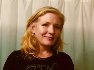 CoraElaine, a blonde woman, looks at the camera while wearing a black t-shirt against a light-colored curtain background.