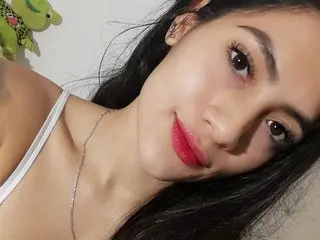 Young woman with long dark hair and red lipstick wearing a white tank top in a bright indoor setting.