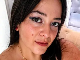 Close-up portrait of a woman with dark hair and winged eyeliner wearing a gold necklace in a bright setting.