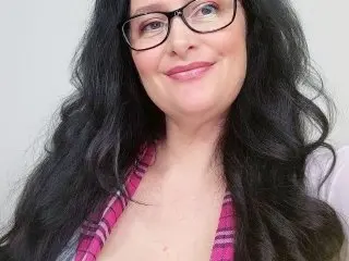 CumForJen smiles while wearing black glasses and a pink and black plaid shirt, showing her long, dark wavy hair.