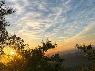 A wide landscape shot of a sunset or sunrise over a distant mountain range, with bright light filtering through dark tree silhouettes in the foreground.
