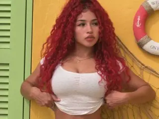 Young woman with long curly red hair wearing a white crop top against a bright yellow beach wall.