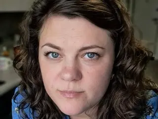 CurlySueSweet looks directly at the camera with her long, dark curly hair framing her face in a close-up headshot.