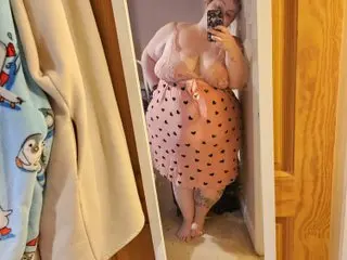 CurvesWithBBW is a curvy woman with blonde hair wearing a pink heart-patterned dress, posing for a mirror selfie in a doorway.