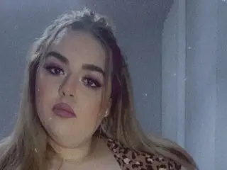 CurvyDollieMae poses with her long brown hair down, wearing a leopard print top against a plain wall.