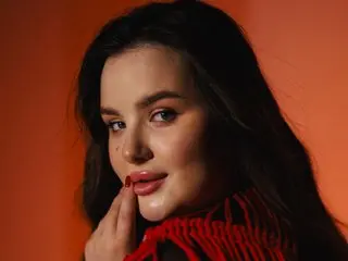 CuteeAmber poses with a soft smile, showing her long dark hair and red top against a warm orange background.