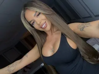 Smiling brunette woman in a black tank top with tattoos posing indoors for a casual selfie.