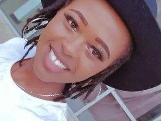 Smiling Black woman with dreadlocks and black hat wearing a white top in bright outdoor lighting.