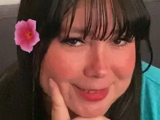 Smiling woman with a pink flower in her hair wearing a teal shirt in a cozy indoor setting.