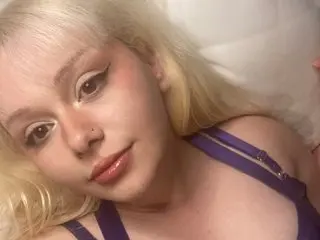Cyber_Melanie poses close-up with her blonde hair, wearing a purple strap, looking directly at the camera.