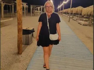 DARLEENEx stands on a wooden boardwalk wearing a short black dress, looking at the camera with blonde hair and glasses.