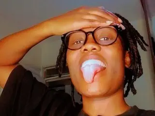 Young woman with dreadlocks and glasses wearing a black shirt, making a playful expression indoors.
