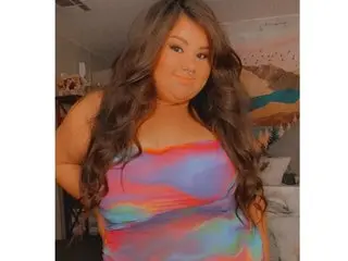 DaniDollxxxx with long brown hair wearing a colorful tie-dye tank top, posing or looking at the camera in a room.