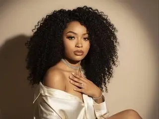 Daniela_Brooks poses with her long, curly hair or black hair, wearing a white silk top that slips off one shoulder, showing her neck and collarbone.