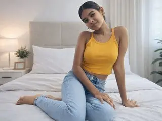 Young woman in a yellow tank top and blue jeans sitting relaxed on a white bed in a bright bedroom.
