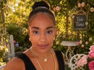 Daniella_Ruby poses with her hair in a bun, wearing a black tank top against a soft, leafy garden background.