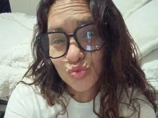 DanielleDeblasio with long wavy hair, wearing a white t-shirt and black glasses, looking into the camera with a pouting expression.