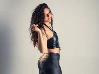 Dannaloves poses in profile, wearing a black leather bra and matching bottoms, showing her long curly dark hair against a plain light background.