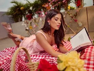 DannyRoge poses outdoors in a pink top, lying on a red and white checkered blanket while holding an open book.