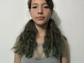 Dante_Katty poses against a white wall, wearing a grey tank top that shows her chest and cleavage, with her long, wavy dark hair falling over her shoulders.