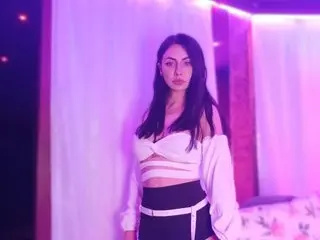 DanyelleVee poses in a white off-the-shoulder top with her long dark hair down against a purple lit background.