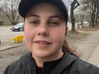 DariaCalm looks at the camera with a slight smile, wearing a black baseball cap and a black jacket against an outdoor background.