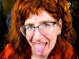 DariaDinkley, a redheaded woman with bangs and glasses, licks her lips while looking at the camera.