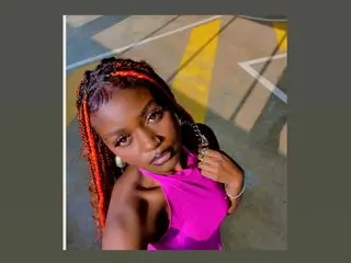 DarkAngelMuseX poses outdoors at night wearing a bright pink halter top with her long red and black braids falling over her shoulders.