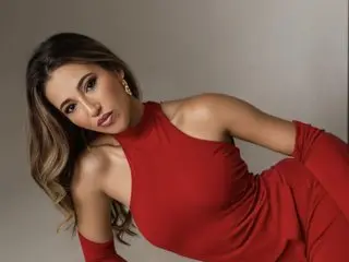 DeeaDivine poses in a red sleeveless dress, letting her long brown hair fall over her shoulder against a plain grey background.