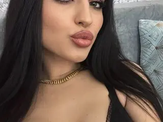DeeaPassion poses with long black hair and a pouty lip, wearing a black top and a gold chain necklace.