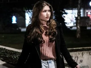 DeliciousFox poses outdoors wearing a pink ribbed sweater and a black jacket with her long brown hair down.