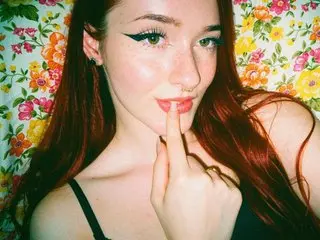 Redheaded woman with winged eyeliner posing playfully against a vintage floral pattern background.