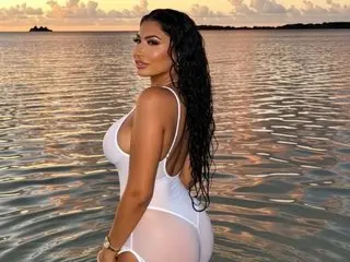 Denise_Taylor poses in a white bodysuit against a sunset ocean background, showing her long dark hair and bare back.