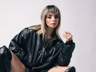 Diavalo poses in a black leather jacket, looking at the camera with her blonde hair styled in a blunt fringe.
