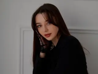 Dinara_Betri poses with her hand near her face, wearing a black top with her long dark hair styled down against a plain white wall.