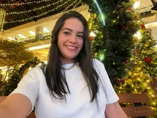 DiosaGrecia18 smiles while posing in a white t-shirt with her long dark hair down in a warm, indoor setting.