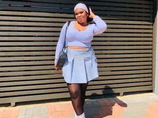 DirtyMelo poses wearing a light blue long-sleeve crop top and a matching short skirt, with her hair pulled back against a slatted wall background.