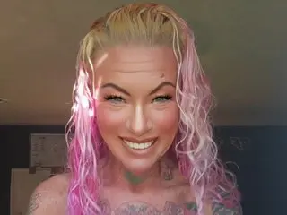 Smiling woman with wavy pink and blonde hair and tattoos in a bright indoor setting.