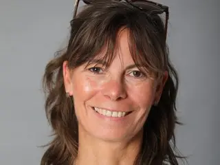Dixie_Milf smiling head-on with her brown hair styled with bangs and sunglasses resting on top of her head against a plain grey background.