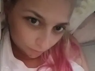 DjLilPuff looks into the camera with pink hair, showing her face and shoulders while lying down.