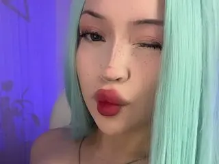 DollWett poses with her light blue hair framing her face, showing her eyes and lips against a purple background.
