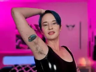 DominaDentata poses with one arm raised, showing her armpit and wearing a black shiny latex top against a pink background.