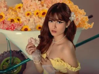 DulceBenett poses in a yellow lace bra with her long brown hair and white flower accessory, surrounded by pink and yellow flowers.