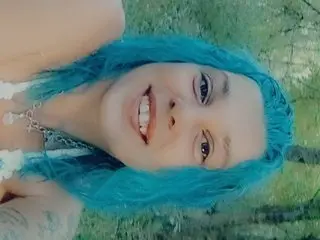 Dwwylin shows her face and blue hair while looking at the camera in a close-up shot.